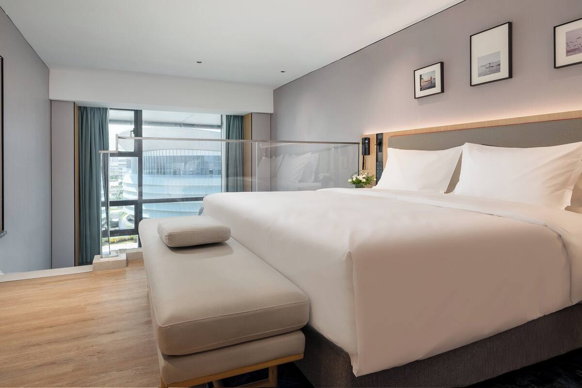 A modern hotel room with a large bed, a bench, a TV, and a view of the city through large windows.
