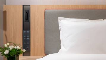 Quarto de hotel com parede de painel de madeira, painel de controle, cabeceira cinza, travesseiro branco e um vaso com flores brancas.