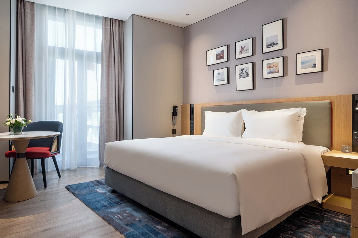 A modern hotel room with a large bed, a desk with a chair, a TV, and framed pictures on the wall.