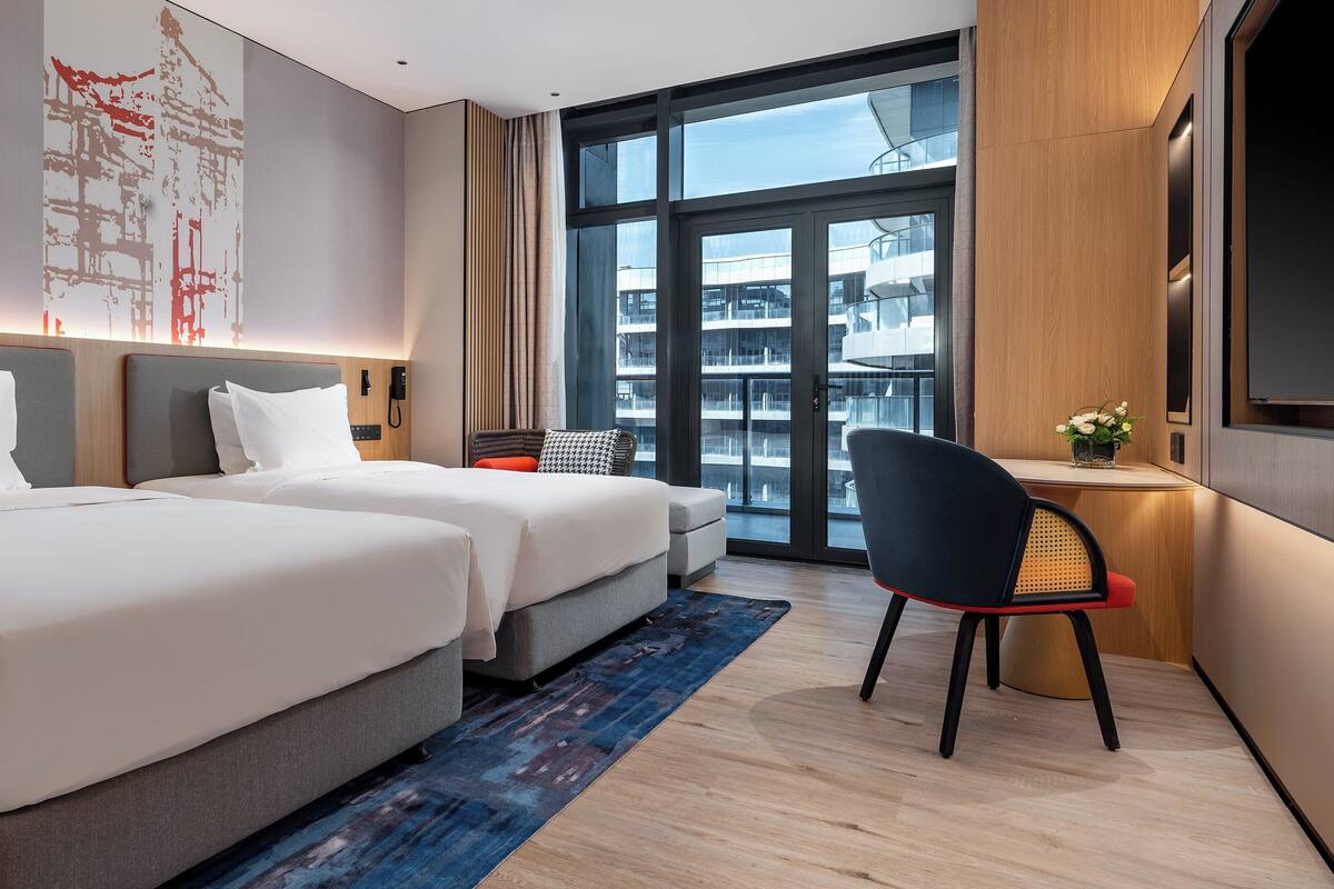 A hotel room with two beds, a desk, a chair, a large window, and a view of the city.