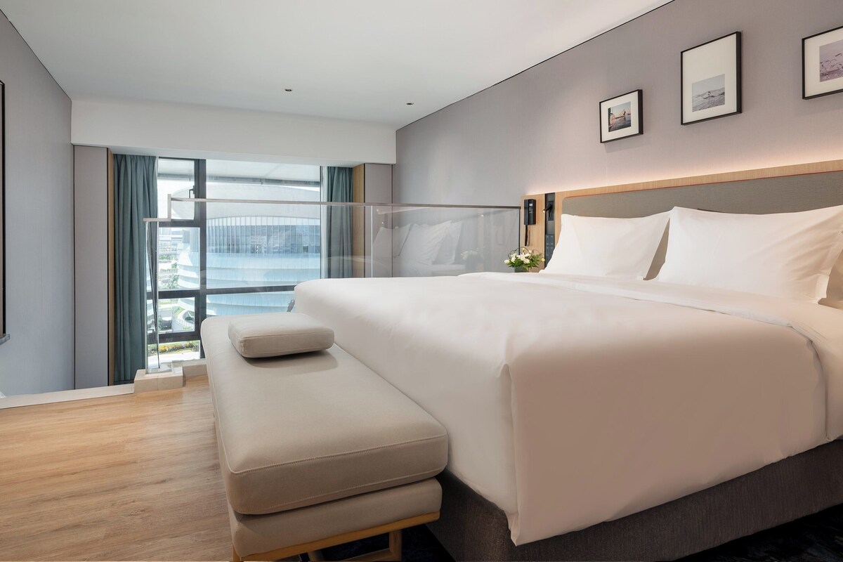 A modern hotel room with a large bed, a bench, a TV, and a view of the city through large windows.