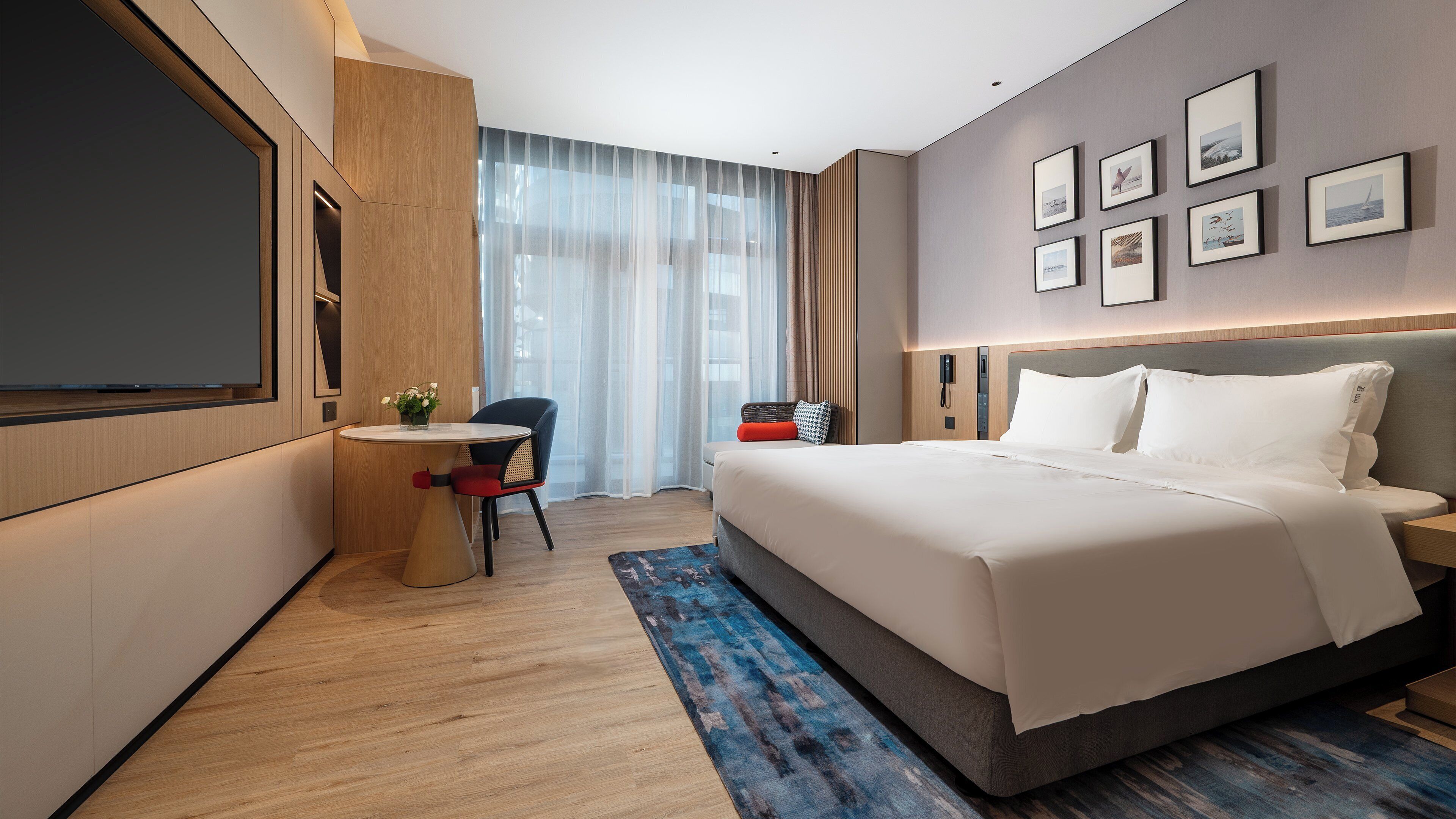 Quarto de hotel moderno com uma cama grande, uma escrivaninha com cadeira, televisão de tela plana e uma grande janela com cortinas finas.