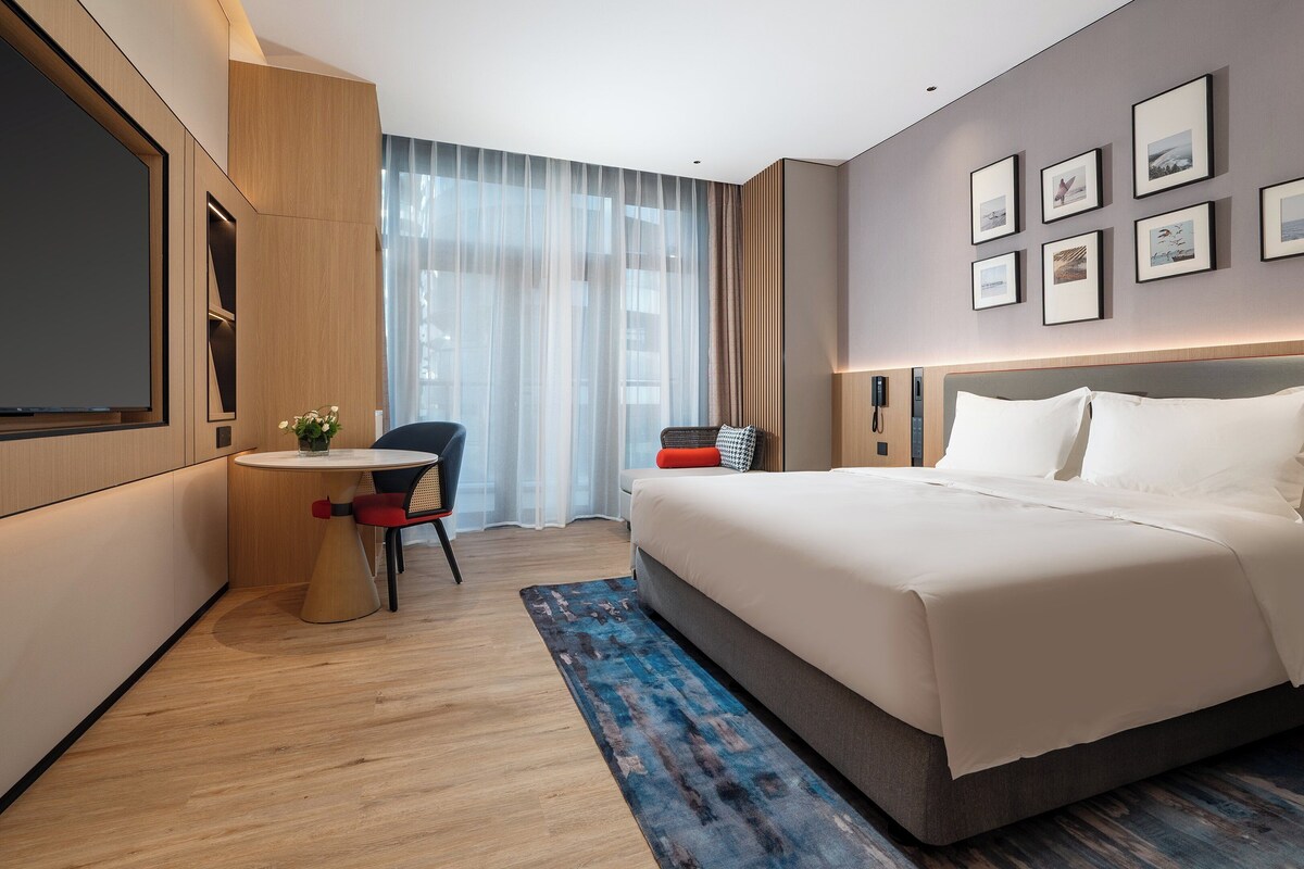 A modern hotel room with a large bed, a desk with a chair, a flat-screen TV, and a large window with sheer curtains.