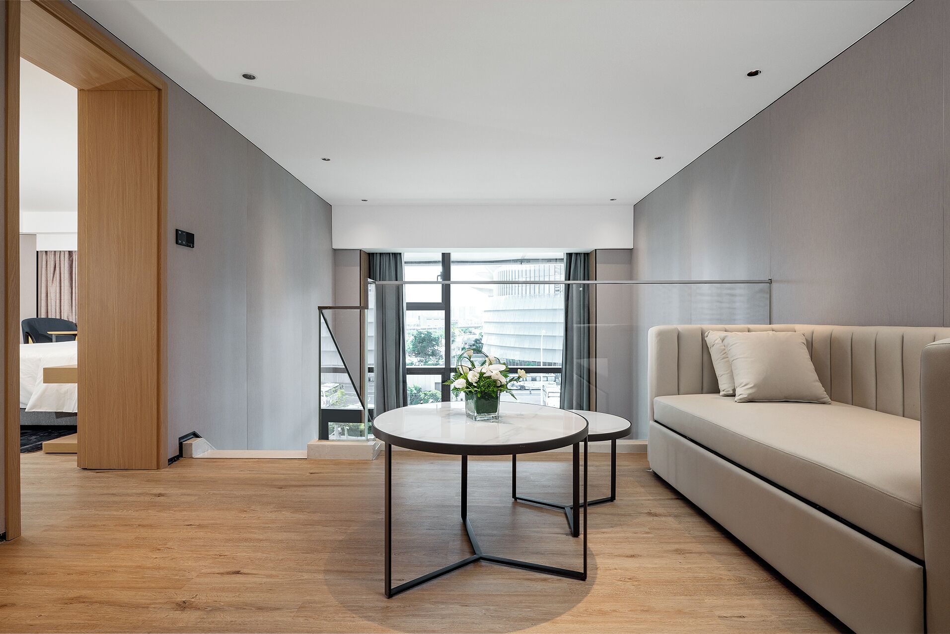A modern hotel room with a sofa, a small round table, and a view of the city through large windows.
