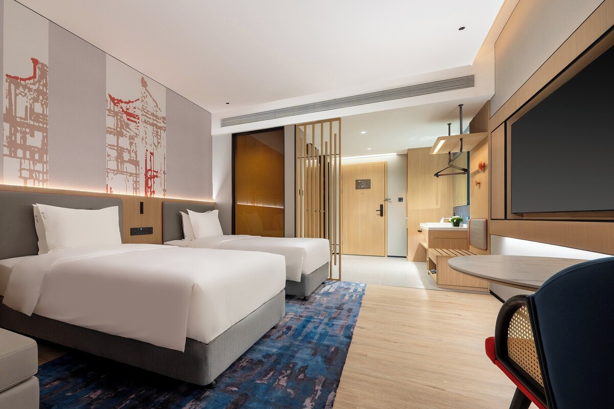 A modern hotel room with two beds, a desk, and a bathroom.