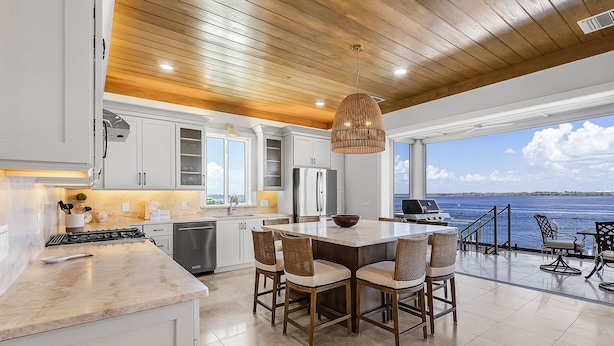 A modern kitchen with white cabinets, a large island, and a dining area with wicker chairs. - 319 Bay and Beach Blessing: Bay Front Pool Home w/ Gulf Views (Fort Myers Beach)