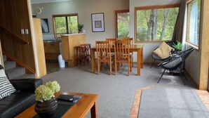A spacious living area with a wooden dining table and chairs, a sofa, and a coffee table with a vase. There are large windows providing natural light and views of the outdoors. - Pukawa Villas (Formerly Oreti Village Resort) (Pukawa)