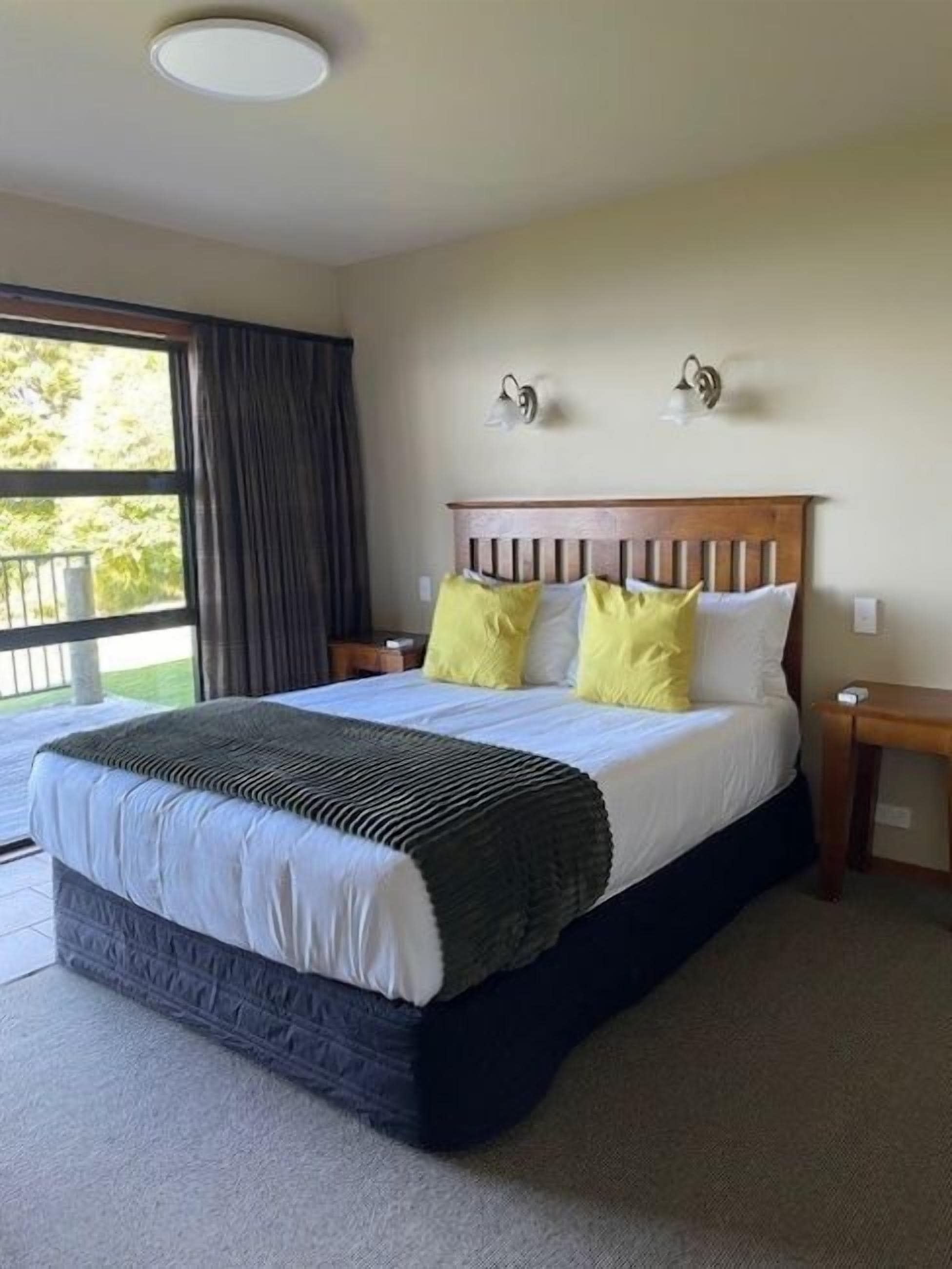 A bedroom with a large bed, two bedside lamps, a window with curtains, and a view of greenery outside.