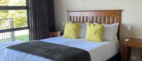 A bedroom with a large bed, two bedside lamps, a window with curtains, and a view of greenery outside.