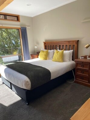 A bedroom with a bed, wooden headboard, bedside tables, and a view of trees through a window. - Pukawa Villas (Formerly Oreti Village Resort) (Pukawa)