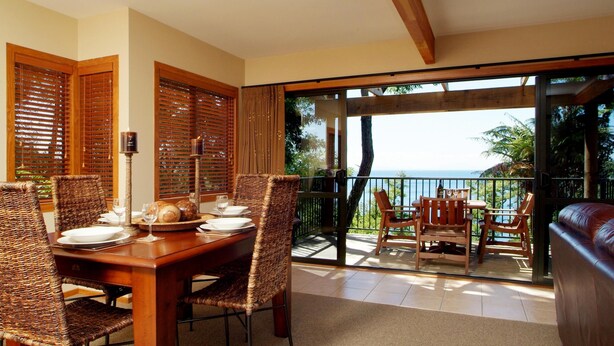 A dining area with a wooden table and wicker chairs, a balcony with a view of the sea, and a leather sofa. - Pukawa Villas (Formerly Oreti Village Resort) (Pukawa)