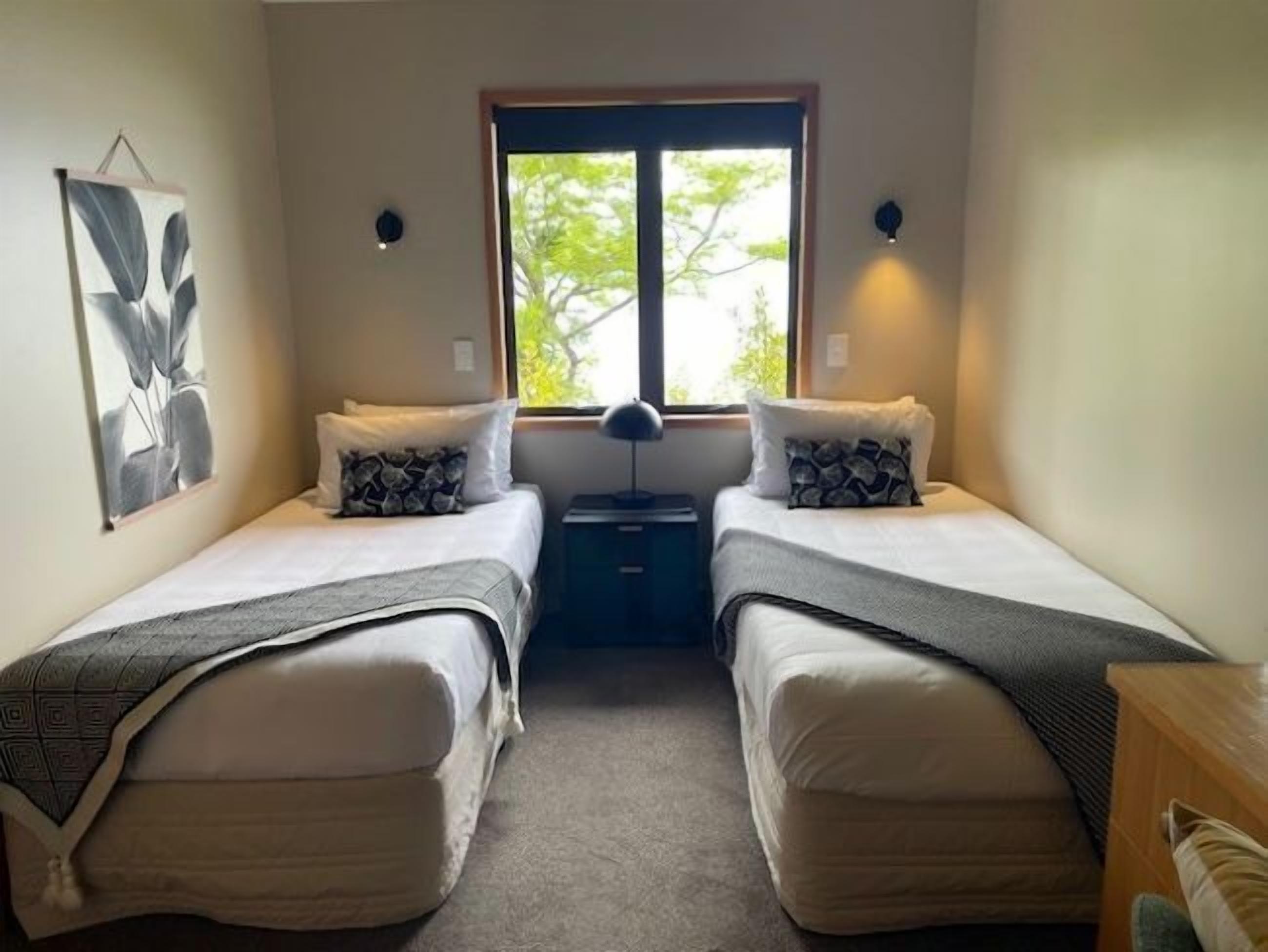 Une chambre avec deux lits simples, une table de chevet, une lampe et une fenêtre donnant sur des arbres.