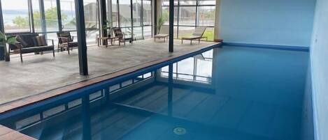 En inomhuspool med fri utsikt mot omgivningen, ett avslappningsområde med fåtöljer och ett betongtak.