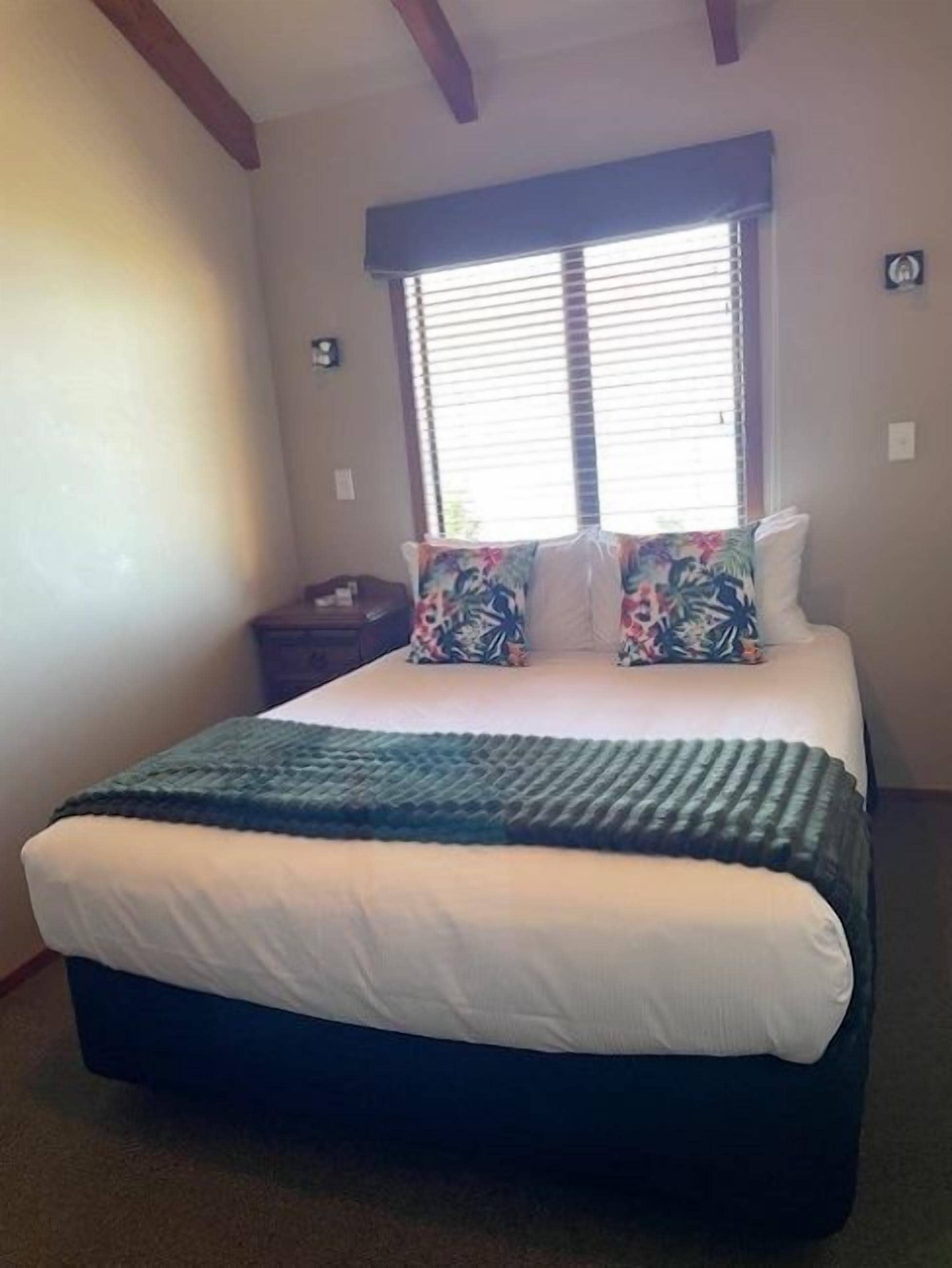 A bedroom with a bed, a window with blinds, a nightstand, and a wall clock.