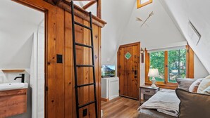 A bedroom with a wooden wardrobe, a bed with pillows, a nightstand with a lamp, and a window with a view of trees. - Timber Lodge Cabins 25 (Colorado Springs)