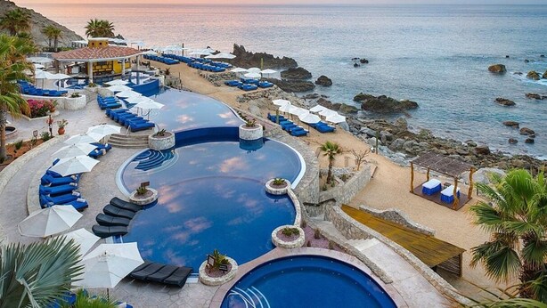 A resort pool with blue lounge chairs and umbrellas, surrounded by a rocky coastline.