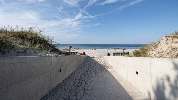 Ein Betonweg führt zu einem Sandstrand, flankiert von Dünen.