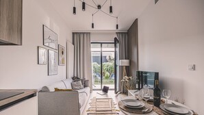 Ein modernes Wohnzimmer mit einem grauen Sofa, einem Esstisch für zwei Personen und einem großen Fenster mit Blick auf Grünflächen.