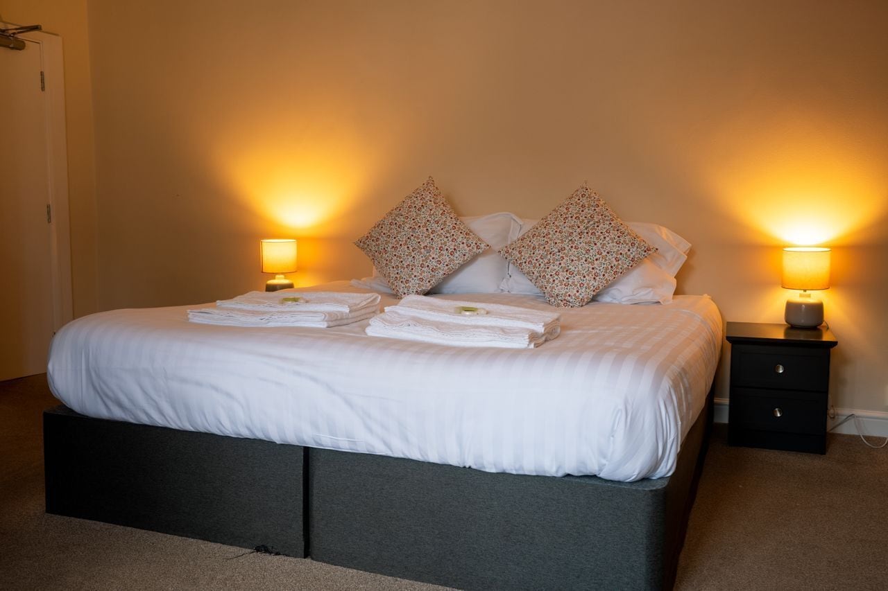 A neatly made bed with white linens and floral-patterned pillows, flanked by two bedside lamps on a dark nightstand.