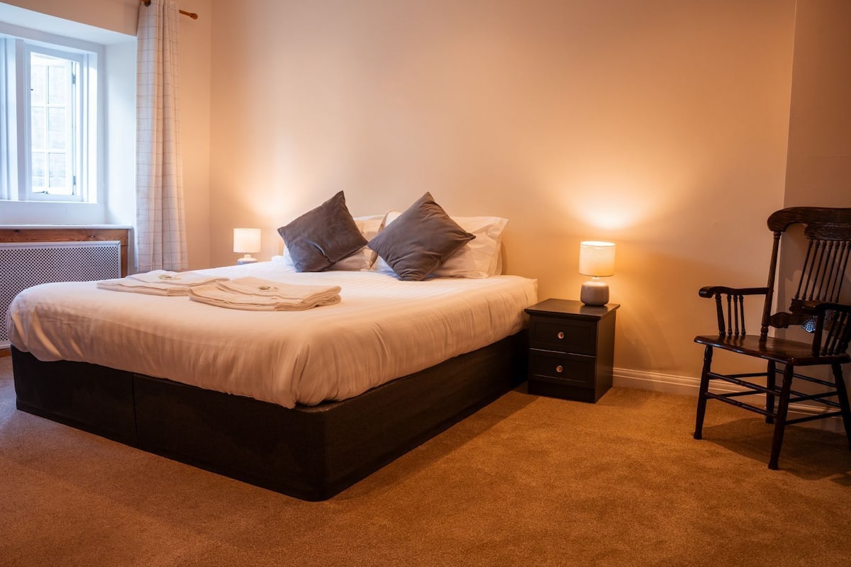 A neatly made bed with pillows, a bedside table with a lamp, and a chair in a well-lit room with a radiator and a window.