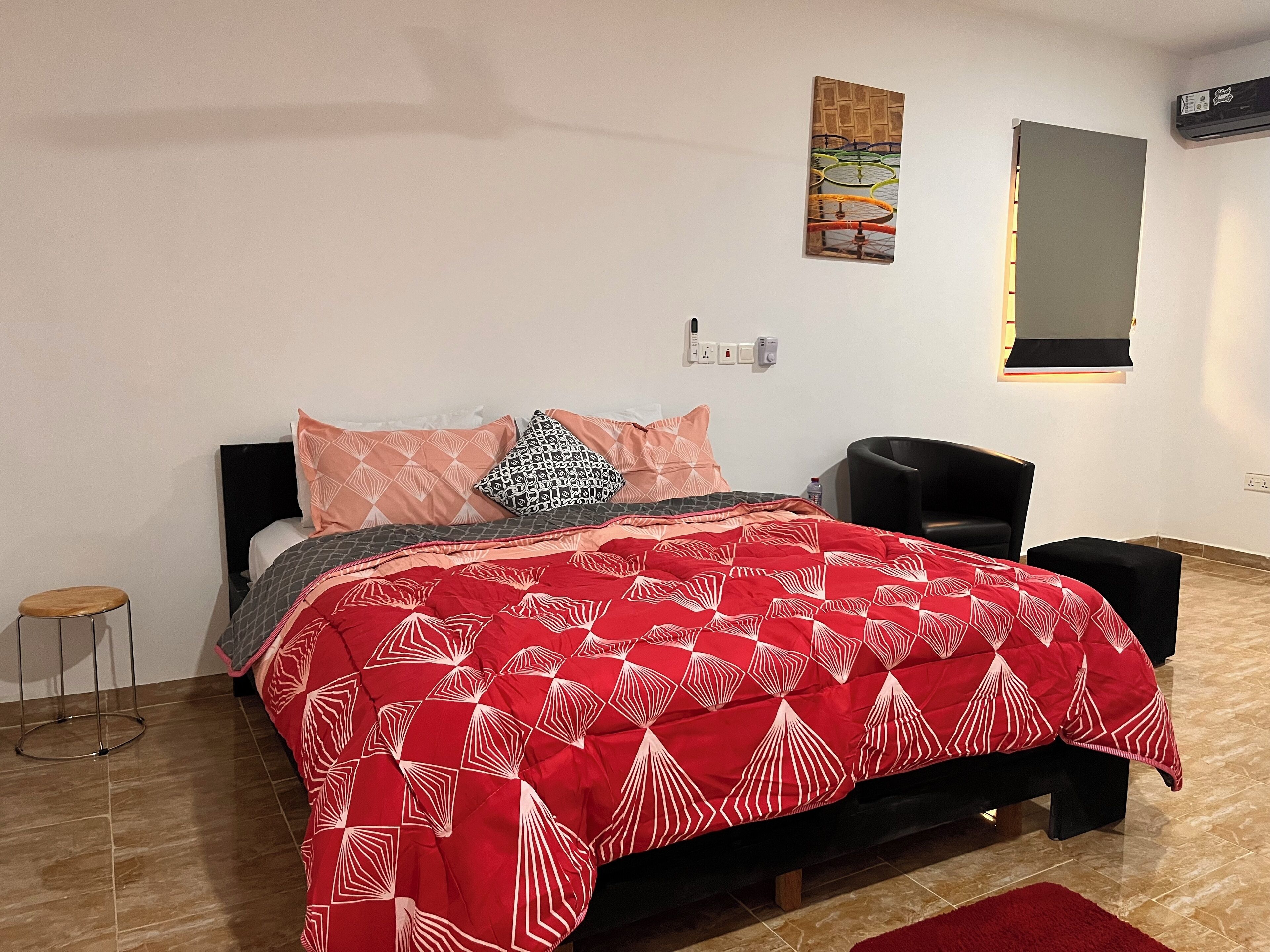 Un dormitorio ordenado con cama, una silla, un taburete pequeño y un aire acondicionado instalado en la pared.