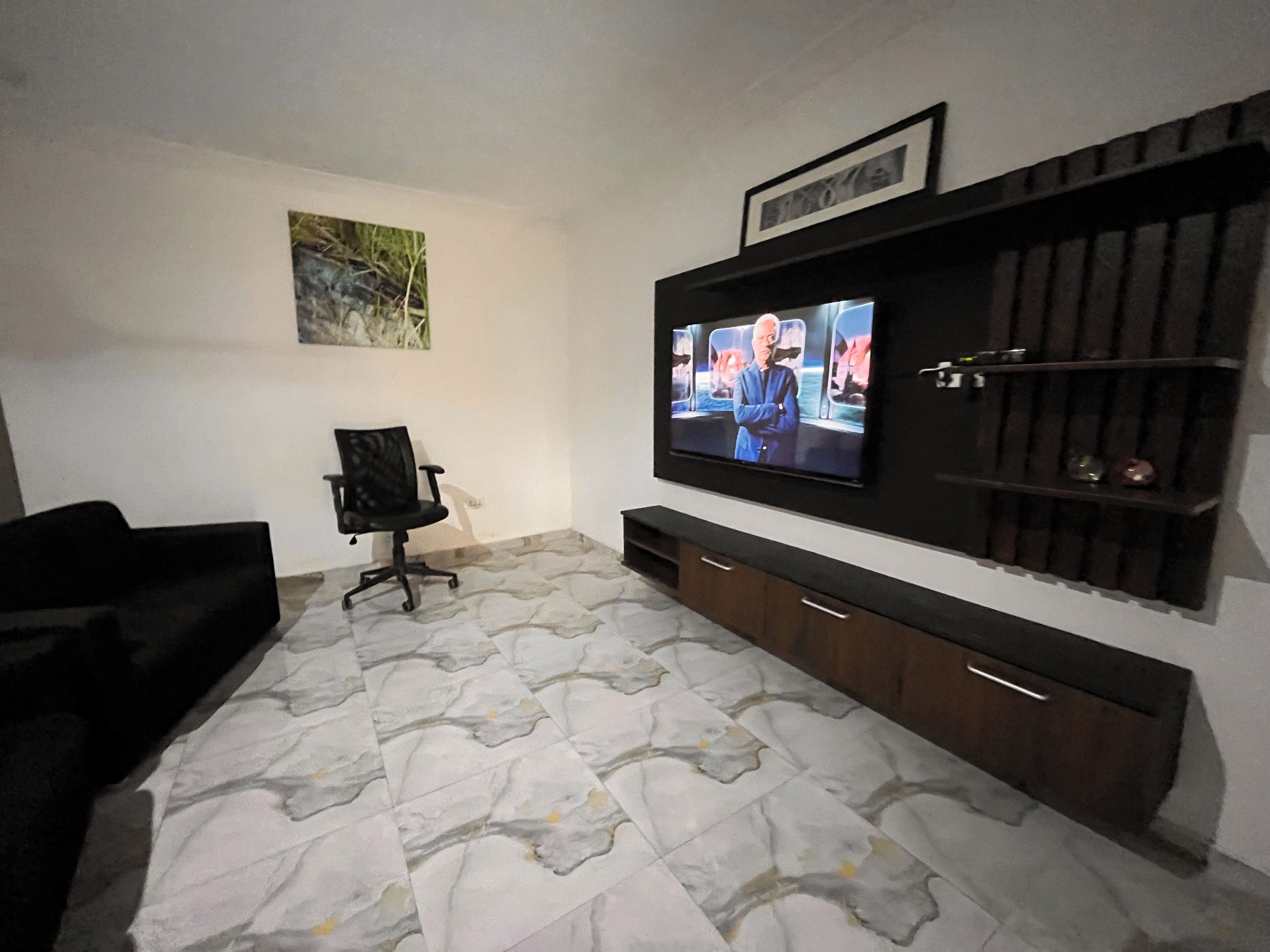 A living room with a flat-screen TV mounted on the wall, a black office chair, and a patterned floor.