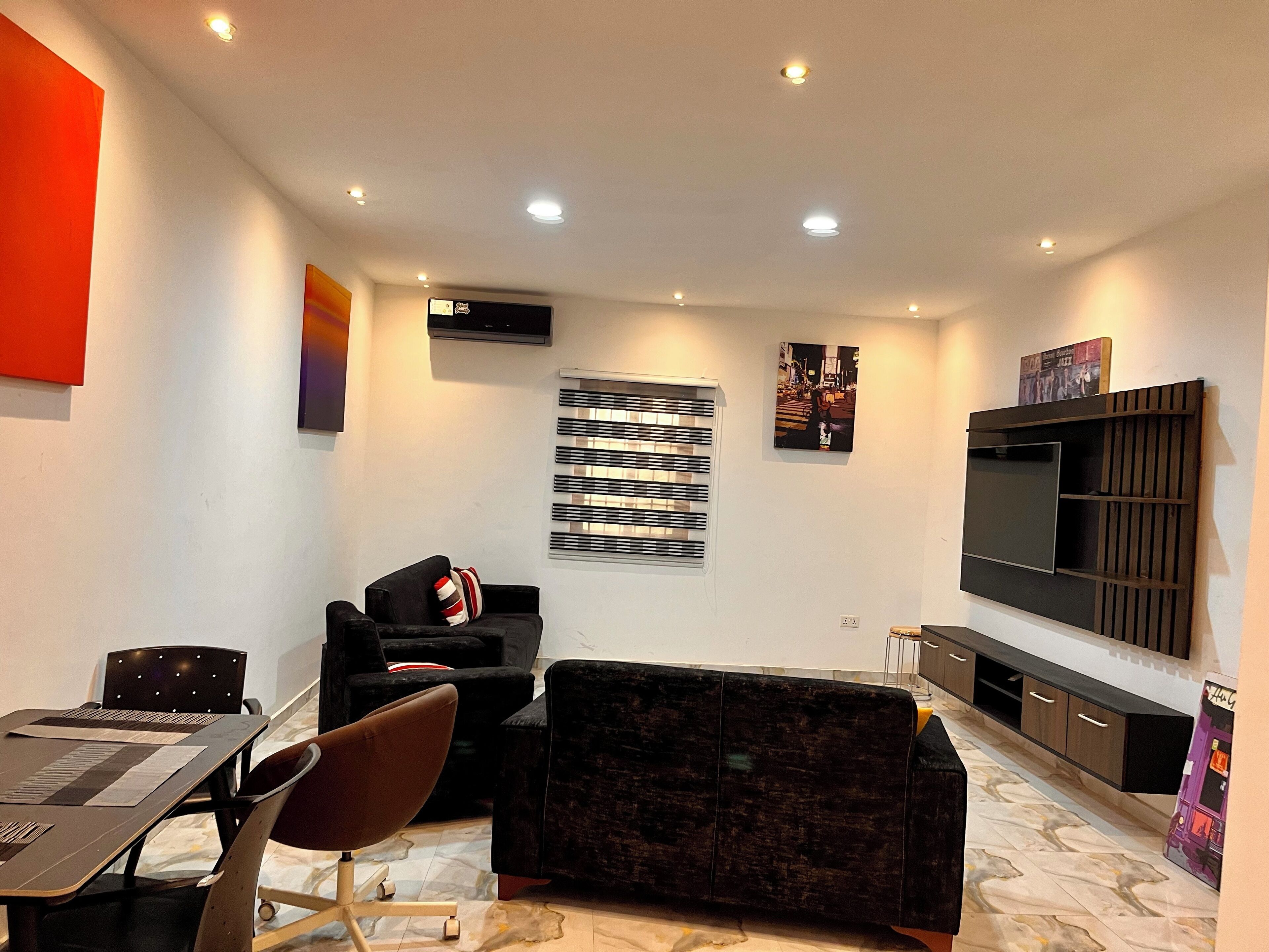 Una sala de estar moderna con un sofá, una mesa de comedor con sillas, un televisor de pantalla plana montado en la pared y una ventana con persianas.