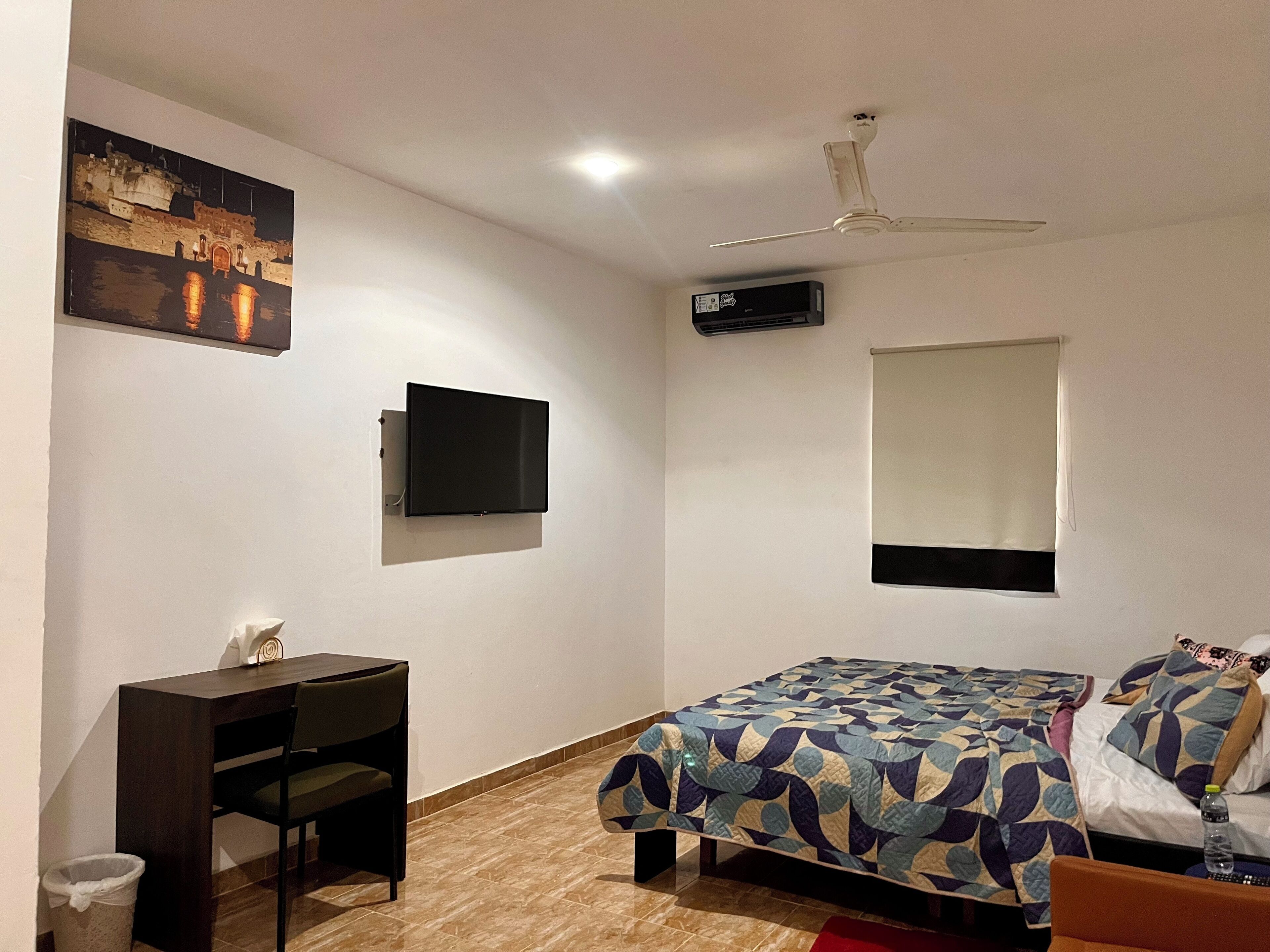 A bedroom with a bed, a desk, a chair, a TV, a ceiling fan, and a window.