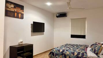 A bedroom with a bed, a desk, a chair, a TV, a ceiling fan, and a window.