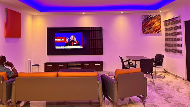 A modern living room with a flat-screen TV mounted on the wall, a sofa, a dining table with chairs, and a decorative wall piece. - ESPADA LODGE Studio 3.3 (Somanya)