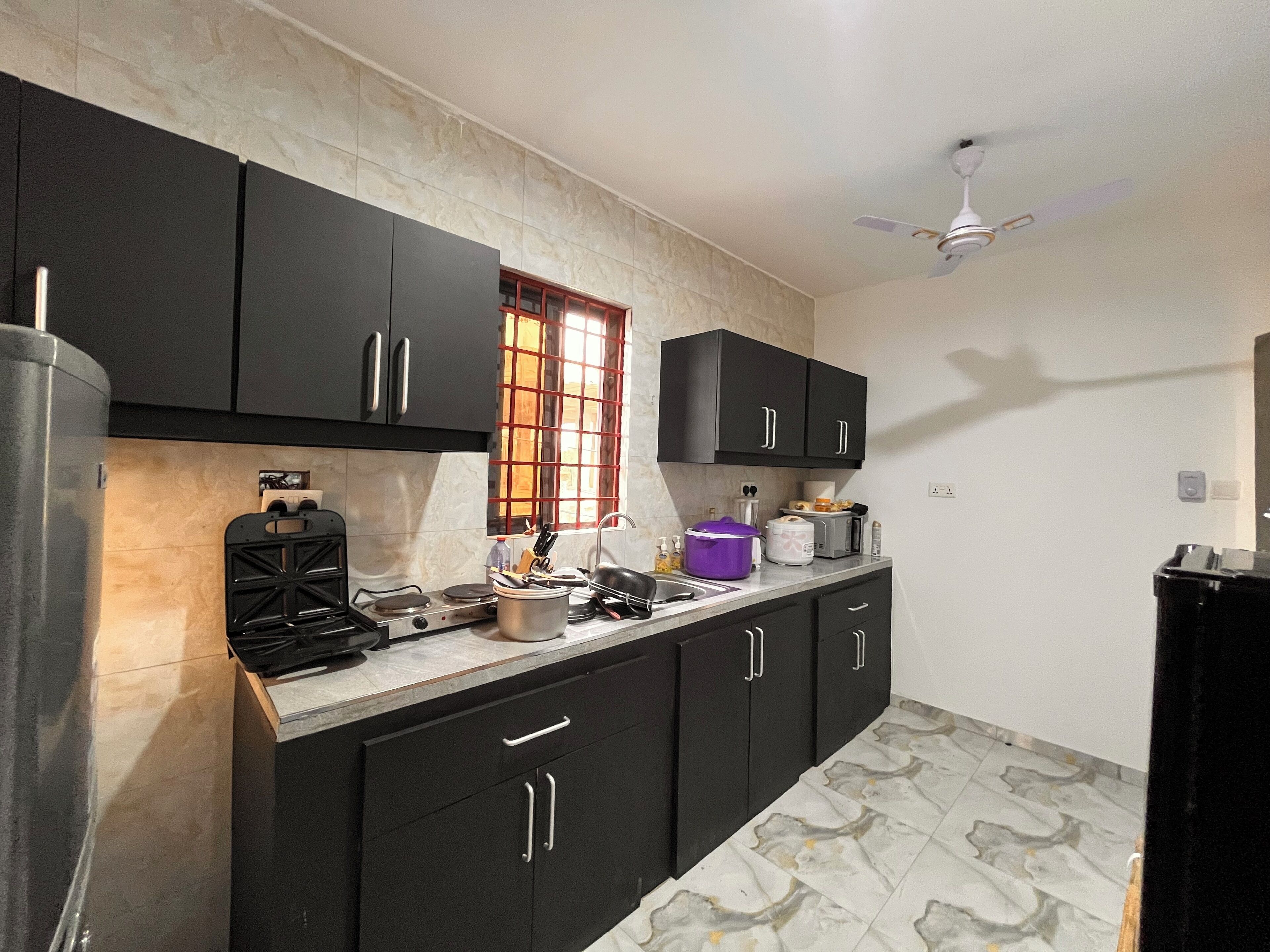 A kitchen with black cabinets, a stove, a window with red bars, and a ceiling fan.