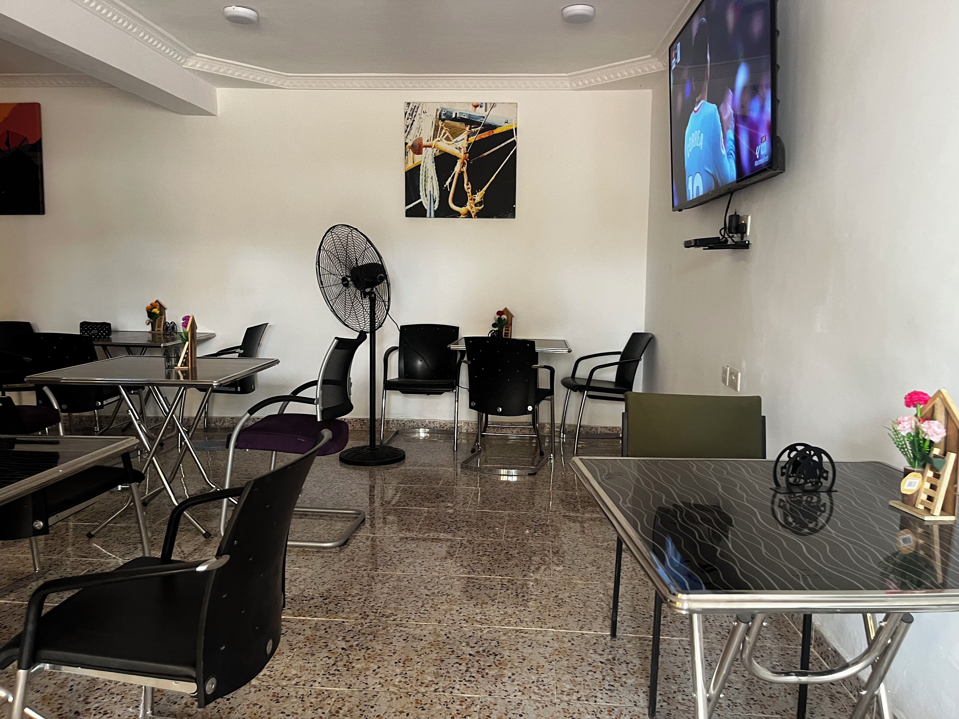 A conference room with tables and chairs, a standing fan, a TV on the wall, and a painting.