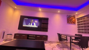 A modern living room with a flat-screen TV mounted on the wall, a dark wooden coffee table, a glass-top dining table with chairs, and a patterned floor. - ESPADA LODGE Studio 3.3 (Somanya)