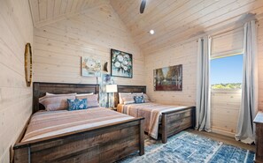 A bedroom with two queen beds, wooden walls, a large window with curtains, and artwork on the walls.
