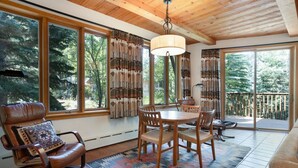 A cozy room with a wooden ceiling, large windows with curtains, a dining table with chairs, a leather chair with a pillow, and a view of trees outside. - Exclusive Cascade Home - Close to Cascade Lift (Vail)