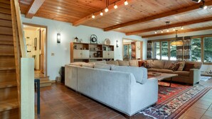 A spacious living room with a large sectional sofa, a smaller sofa, and a coffee table. There is a bookshelf with various items and a clock on the wall. The room has wooden flooring and a large window with a view of trees. - Exclusive Cascade Home - Close to Cascade Lift (Vail)