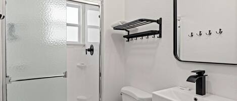 Un baño con inodoro y lavamanos blancos, un grifo negro, toallero y puerta de ducha de vidrio.