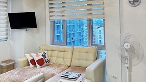 Un salon moderne comprenant un ensemble de canapés, un lustre, un téléviseur à écran plat, un ventilateur sur pied et une fenêtre avec des stores.