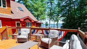 Una terraza de madera con mobiliario de mimbre, una mesa y vistas a un lago y árboles.