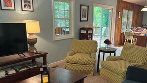 A living room with a TV, a coffee table, a sofa, and patterned chairs. There are framed pictures on the wall and a window with a view of greenery.