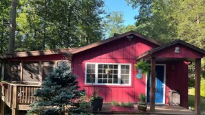 A red cabin with a porch and a small attached shed, surrounded by trees and greenery.