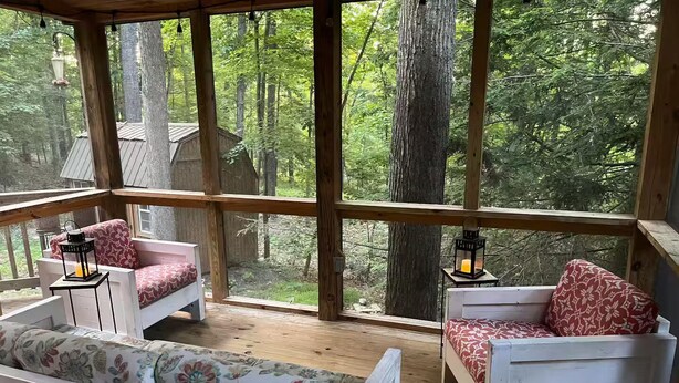 A wooden deck with white furniture and red floral cushions, overlooking a forested area.