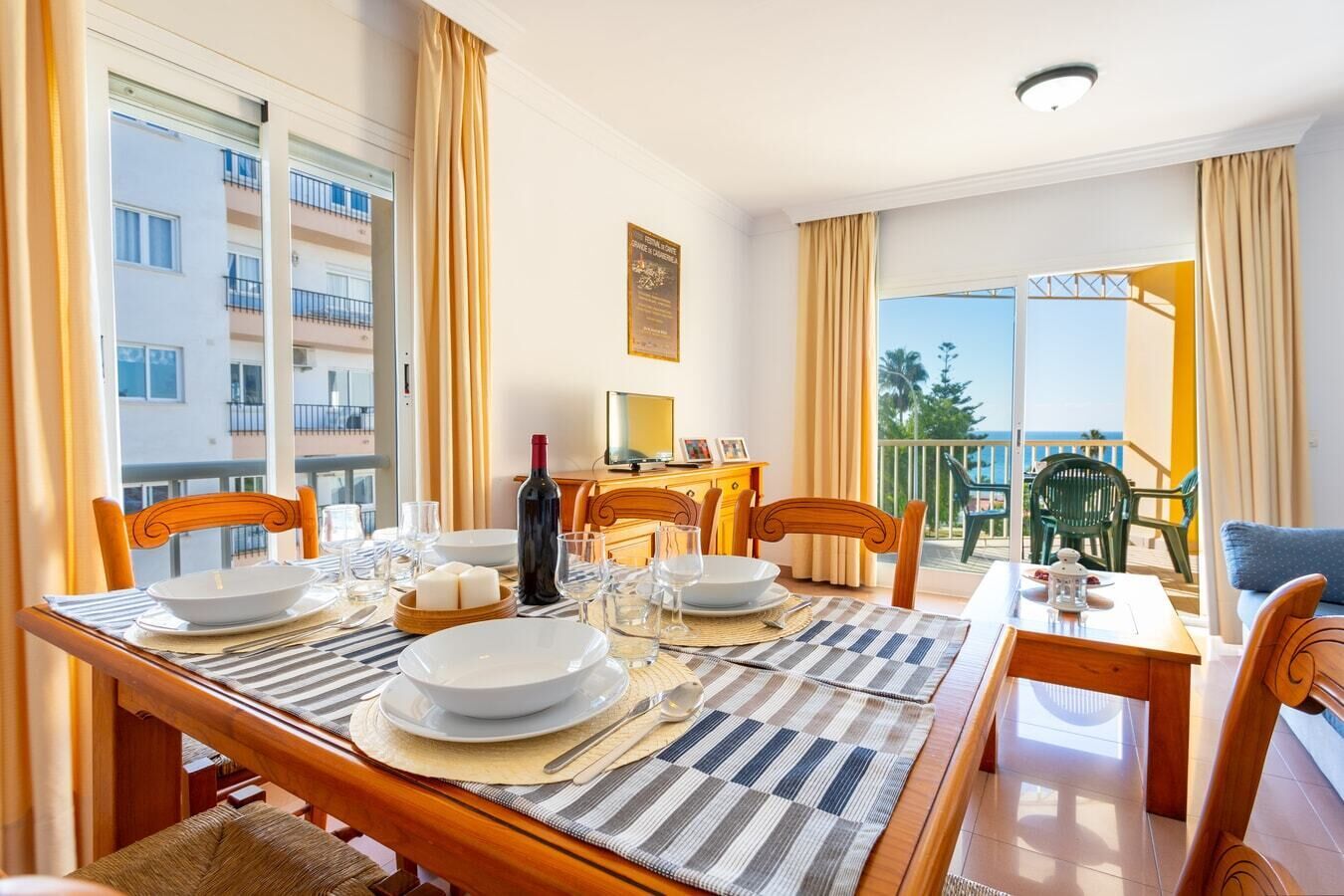 A dining area with a table set for four, a bottle of wine, and a view of the sea through large windows.