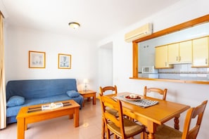 A compact living space with a blue sofa, wooden dining table, and chairs, a small side table, and a kitchen area with cabinets and a microwave.
