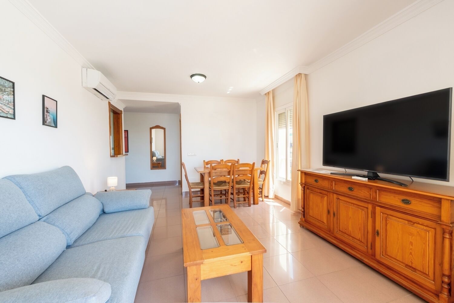 A living room with a sofa, a wooden coffee table, a TV on a wooden cabinet, and a dining area with chairs and a table.
