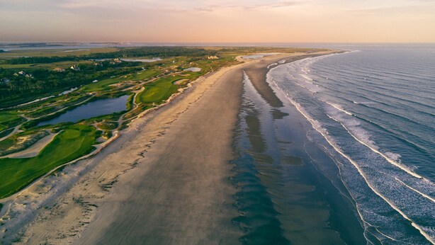 Vista aérea de una zona costera con una extensa playa arenosa, un campo de golf y una masa de agua.