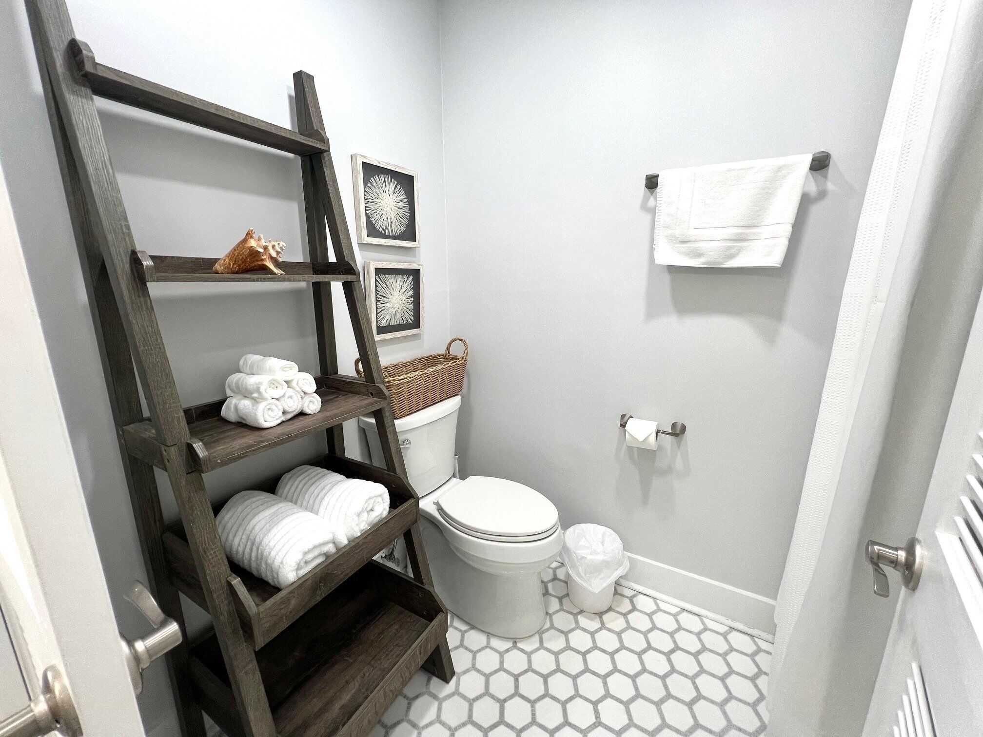A compact bathroom with a wooden shelf holding towels, a toilet, a trash bin, and a wall-mounted towel rack.