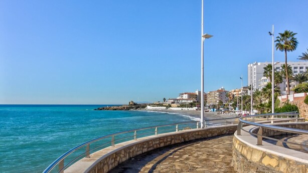 Eine Küstenpromenade mit einem Pflasterweg, einem Metallgeländer und freiem Blick auf das Meer und Gebäude im Hintergrund.