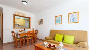 Ein Wohnzimmer mit Esstisch, Holzstühlen, einer Couch mit grünen Kissen und zwei gerahmten Bildern an der Wand.