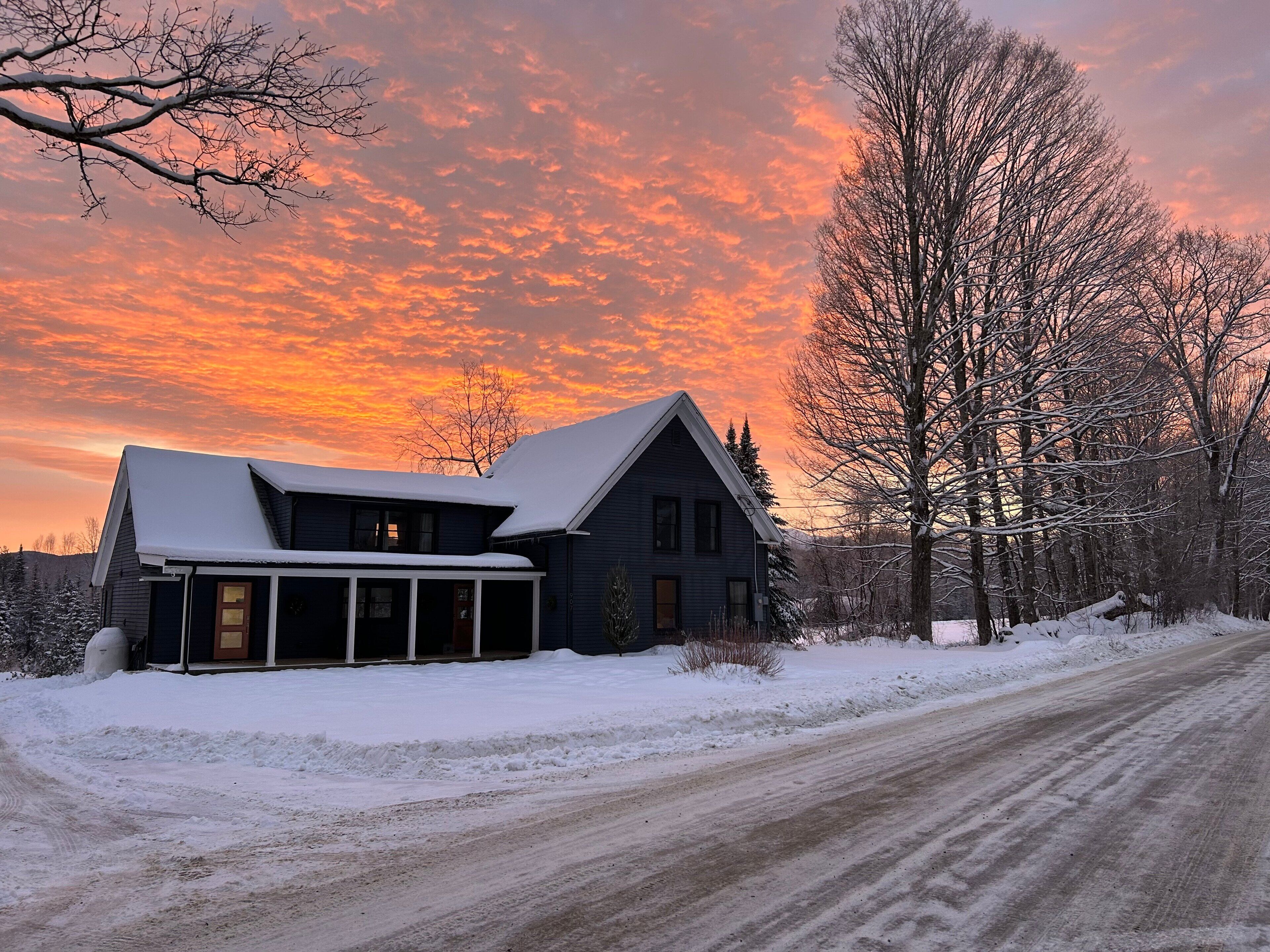 Une maison dont le toit est recouvert de neige, avec un porche, entourée d’un paysage enneigé et d’arbres.